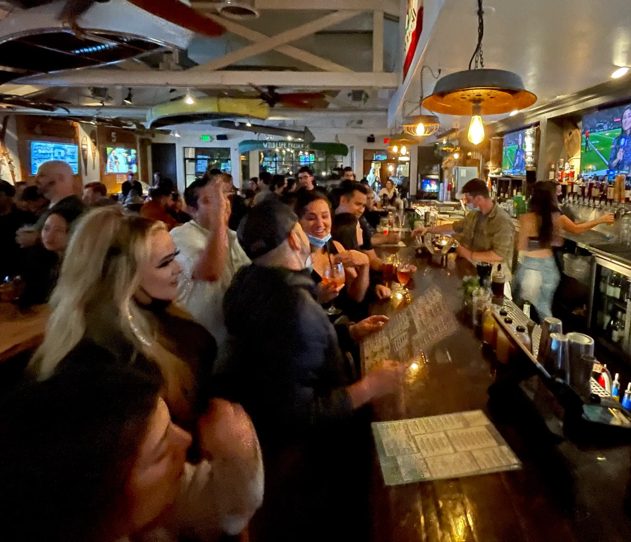 Lively bar scene at Unbearable Skee Lodge with a full crowd at the bar, bartenders pouring drinks, and TVs in the background